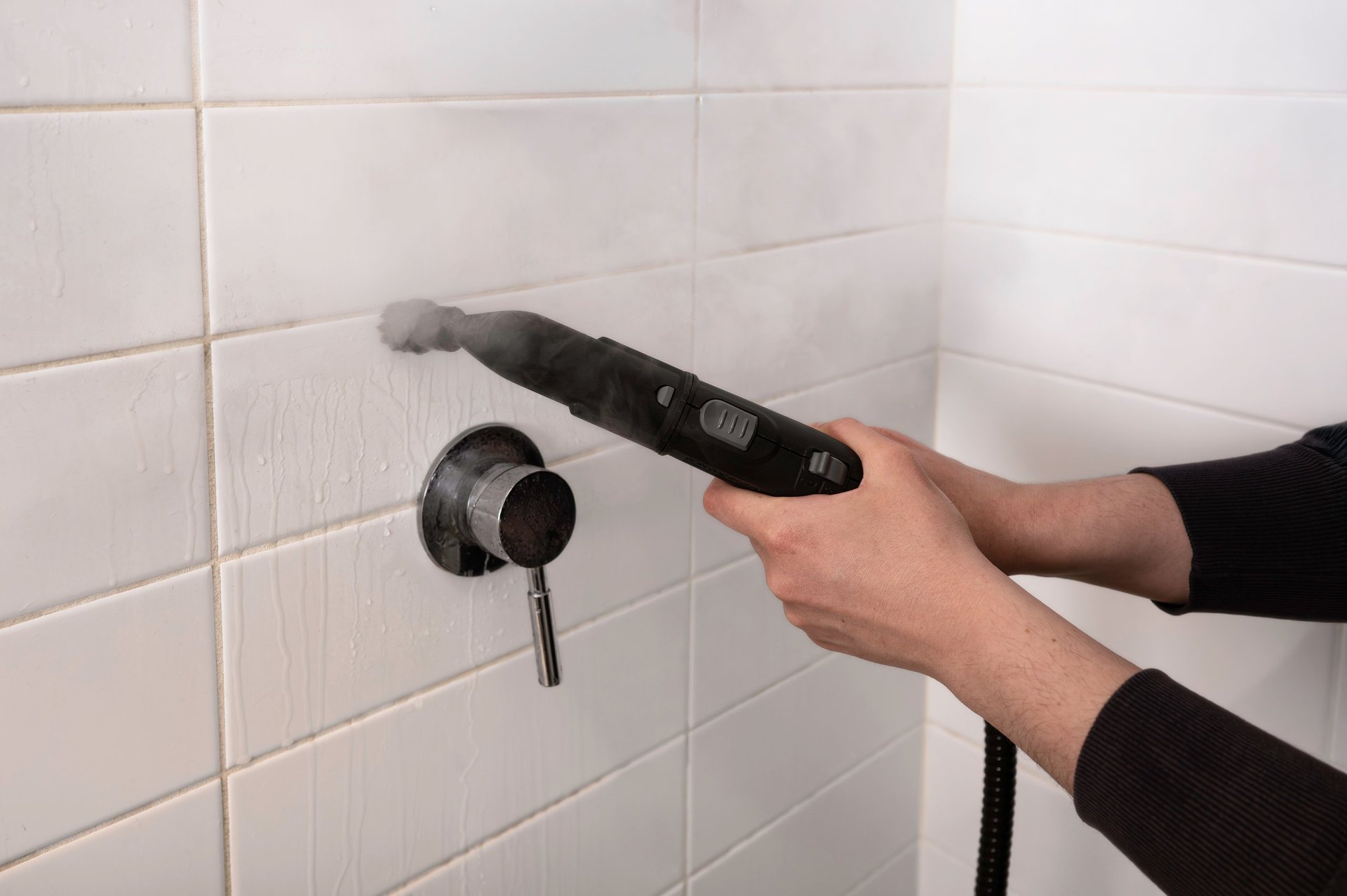Close-up of a person cleaning white bathroom tiles with a handheld steam cleaner