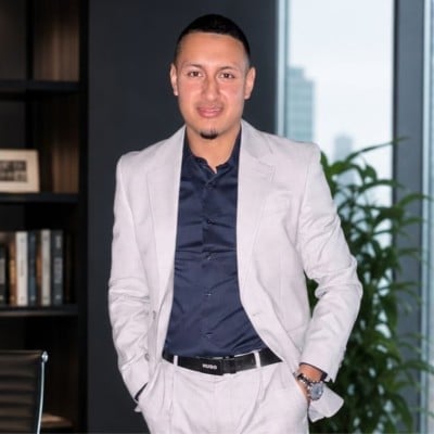 Man in light beige blazer and navy shirt standing in modern office with bookshelves and plant in background