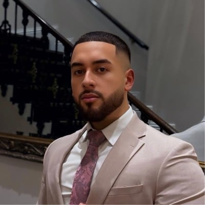 Man with beard wearing beige blazer and burgundy tie, standing indoors near a staircase